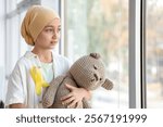 Little girl after chemotherapy with yellow ribbon and toy bear near window. Childhood cancer awareness concept