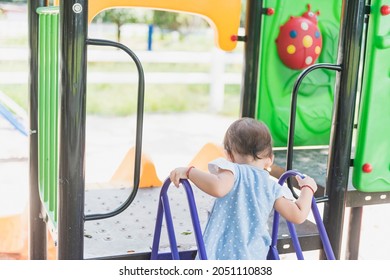 A Little Girl Alone Stood On The Stairs At The Top Of The Slide. Back View, The Concept Of Danger From The Playground.