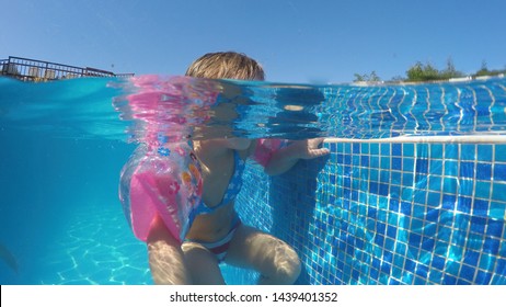 Little Girl With Arm Bands In Pool Makes A Funny Tongue Out Grimaces Toward Camera, Half Underwater Shot