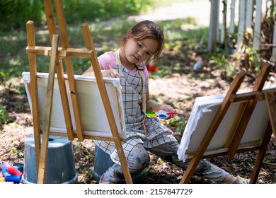 Little Girl Artist Drawing A Picture On Canvas On An Easel In Nature, Cute Little Girl Painting Picture With A Brush And Color In Backyard.  Concept Of Art, Hobby And Relax