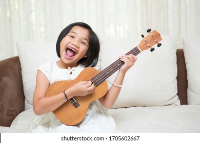 Little Girl Asian Kid Holding Ukulele Or Small Guitar With Four Strings, Smile And Enjoy Playing Music Instrument At Home. Portrait Of Young Girl Musician In Cozy Environment.