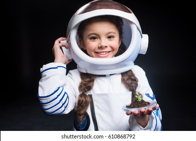 Little girl in astronaut costume and helmet holding fresh plant in soil and smiling at camera isolated on black