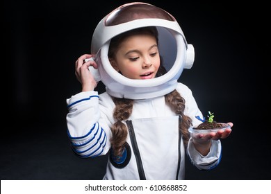 Little Girl In Astronaut Costume And Helmet Holding Fresh Plant In Soil Isolated On Black