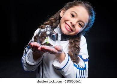 Little girl in astronaut costume holding terrarium jar with plant and smiling at camera isolated on black - Powered by Shutterstock