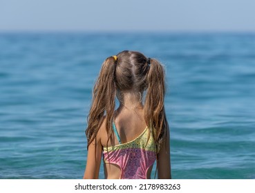 A Little Girl In A Bathing Suit Looks At The Sea. View From The Back.