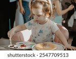 A little girl with a beautiful ponytail sits at the kitchen table and eats pancakes, painting them with edible paints