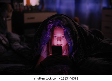 Niña en la cama bajo una manta mirando el smartphone de noche.