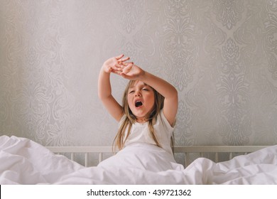 Little girl in the bedroom is sitting on the bed. Little girl is wearing a pajamas and sitting in bed stretching and yawns.
