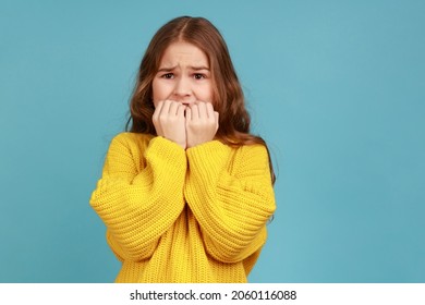 Little girl biting nails with scared big eyes, feeling worried, anxious about childish mistake, wearing yellow casual style sweater. Indoor studio shot isolated on blue background.