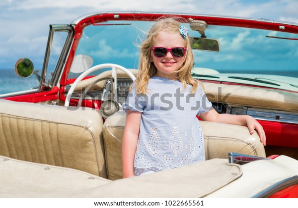 Little girl with blond hair, sunglasses and blue\
blouse is standing inside red retro car cabriolet on the coastline\
of Caribbean sea