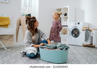 A little girl with blonde hair wearing a dress spends time with her mother in the laundry room, bathroom, a woman sits on the floor sorting clothes, folding laundered items - Powered by Shutterstock