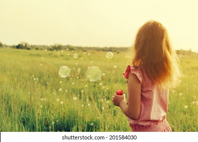 Little girl is blowing a soap bubbles in summer meadow, rear view. With instagram filter.