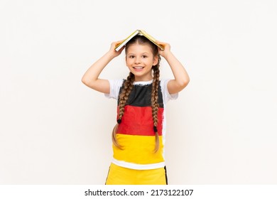 A little girl with a book on her head on a white isolated background. Learning German in the lower grades. A child in a T-shirt with the flag of Germany.