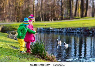 Little Girl And Boy Feeding Ducks And Geese In Park River On Cold Autumn Day. Kids Taking Care Of Animals. Outdoor Fun For Children In Fall Or Winter. Family Day Trip To Birds Zoo.