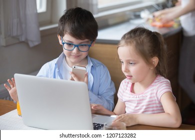 Little Girl And Boy With Gadgets Sitting At The Table At Home. Smart Brother Hold Mobile Phone Using Voice Assistant Sister Surfing Internet Use Computer, New Generation Addicted With Devices Concept