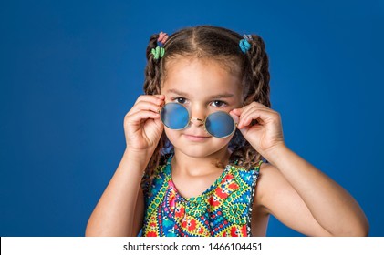 Little Girl With Braids Looking Over Blue Hippie Glasses Sunglasses Isolated On Blue Background