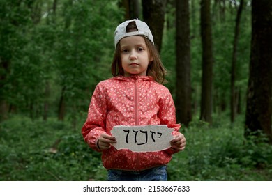 A little girl in a cap stands in a gloomy forest and holds a sign with the inscription in Hebrew remember
