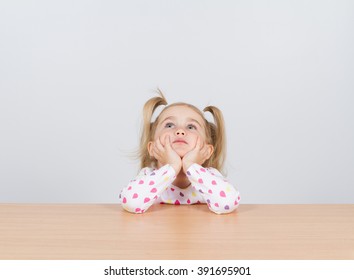 Little girl in casual clothes looking up and thinking. Sitting at the table.