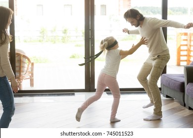 Little Girl Catching Father Playing Hide And Seek, Preschool Blindfolded Daughter Having Fun With Mom And Dad At Home, Parents With Kid Spending Time Together In Living Room On Leisure, Family Games