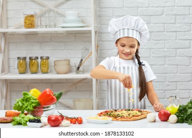 Little Girl In Chef Hat And An Apron Cooking Pizza In The Kitchen. Child Adding Cheese Into Pizza