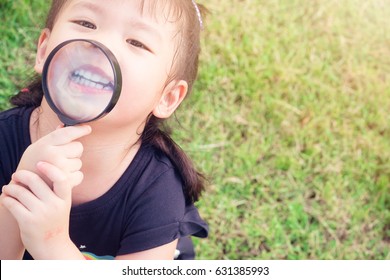 Little Girl Child Holding Magnifying Glass And Showing Front Teeth With Big Smile On Green Grass: Healthy Happy Funny Smiling Face Young Adorable Lovely Female Kid With New Tooth Dental Loss.