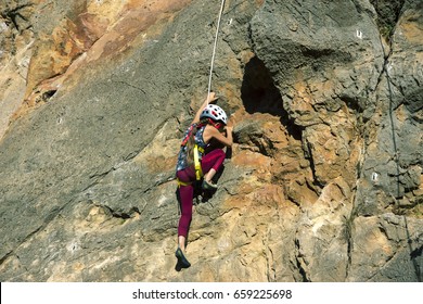 Little Girl Child In Protective Helmet Training Rock Climbing On Natural Outdoor Wall With Belay Rope Fixed To Harness