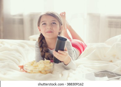 Little Girl Child With Sna?ks And Remote Control Watching Tv Set Lying On Bed In Room. 6 Years Old Child Watching Tv Laying Down On A White Carpet At Home Alone. Cute Little Girl 