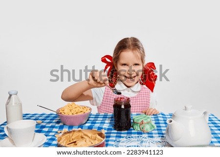 Little girl, child in retro dress eating berry jam, having breakfast against grey studio background. Concept of childhood, game, friendship, activity, leisure time, retro style, fashion.