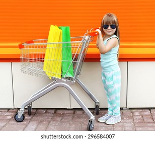 Little Girl Child And Shopping Cart With Bags Against The Colorful Orange Wall