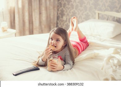 Little Girl Child With Snacks And Remote Control Watching Tv Set Lying On Bed In Room. 6 Years Old Child Watching Tv Laying Down On A White Carpet At Home Alone. Cute Little Girl 