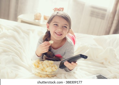 Little Girl Child With Snacks And Remote Control Watching Tv Set Lying On Bed In Room. 6 Years Old Child Watching Tv Laying Down On A White Carpet At Home Alone. Cute Little Girl 