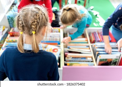 Little Girl Chooses, Takes Book With Fairy Tales From Box In Children's Library.Special Reading Kids Room.Many Shelves With Bright Interesting,educational Books About Animals.