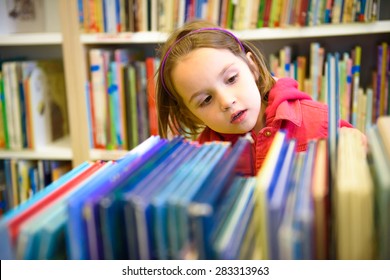 Little Girl Is Choosing A Book In The Library. A Child Is Looking At The Books In The Library Deciding Which One To Take Home. Children Creativity And Imagination.