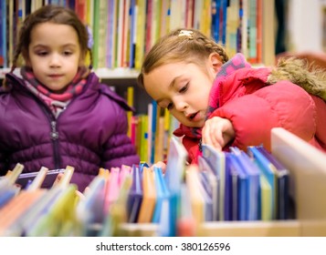 Little Girl Is Choosing A Book In The Library. A Child Is Looking At The Books In The Library Deciding Which One To Take Home. Children Creativity And Imagination.