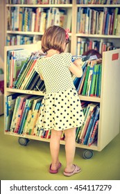 Little Girl Is Choosing A Book In The Library. A Child Is Looking At The Books In The Library Deciding Which One To Take Home. Children Creativity And Imagination.