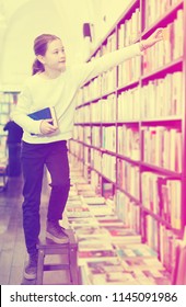  Little Girl Climbing Wooden Stepladder For Taking Book From Tall Bookcase In Shop