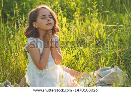Little Girl closed her eyes, praying in a field during beautiful sunset. Hands folded in prayer concept for faith, spirituality and religion. Peace, hope, dreams concept