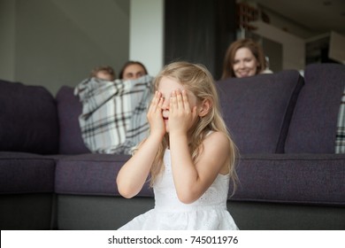 Little girl closing eyes covering face with hands playing hide and seek game with parents and brother hiding behind sofa peeking out in living room, happy family having fun together with kids at home