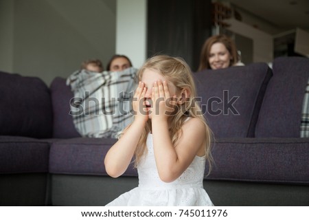 Little girl closing eyes covering face with hands playing hide and seek game with parents and brother hiding behind sofa peeking out in living room, happy family having fun together with kids at home