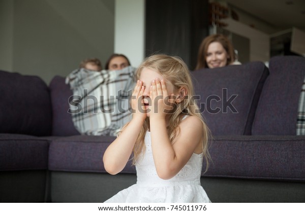 Little girl closing eyes covering face with hands\
playing hide and seek game with parents and brother hiding behind\
sofa peeking out in living room, happy family having fun together\
with kids at home
