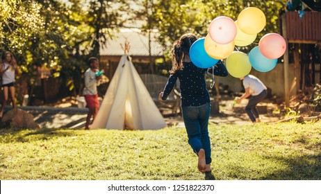 Little Girl With Colorful Balloons Running Towards Kids Playing In Backyard. Children Having Fun Outdoors.