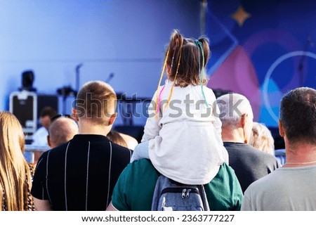 A little girl with colorful pigtails sits on her father's shoulders during a mass outdoor concert. A crowd of people watches a show at a concert venue. Unrecognizable person. Foreground