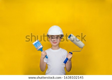 little girl in a construction white helmet holds a paint brush and roller on a yellow background. concept of renovation and construction