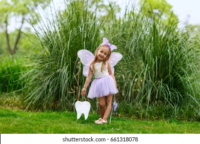 A Little Girl In The Costume Of A Tooth Fairy Stands On The Street And Holds A Bag In The Shape Of A Tooth
