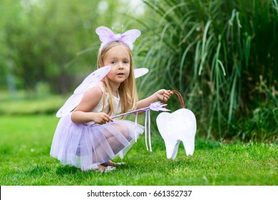 A Little Girl In The Costume Of A Tooth Fairy Sitting In The Park And Holds A Bag In The Shape Of A Tooth