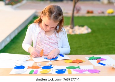 Little Girl Create A Greeting Card Image Of The Jewish Holiday Of Hanukkah. Kid Pastes Stickers - Menorah, Candles, Dreidel, Oil Jar On Paper On Wooden Table. Needlework, Crafts For Children.