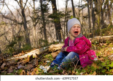 A little girl is crying loudly in the autumn (spring forest); She sits on the grass and fallen leaves; She opens her mouth wide and screams; On the face emotions of despair and resentment