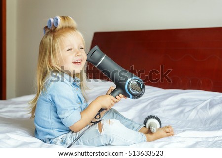 little girl with curlers on her head sitting at home on the bed dries the hair dryer