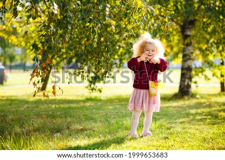 A little girl with curly white hair dressed in a burgundy sweater walks in the park on a sunny day.