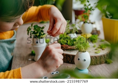 Little girl decorating eggshells with grass with toy stickers eyes on the wooden table at home. Creative fun DIY idea for festive Easter decoration. Selective focus.